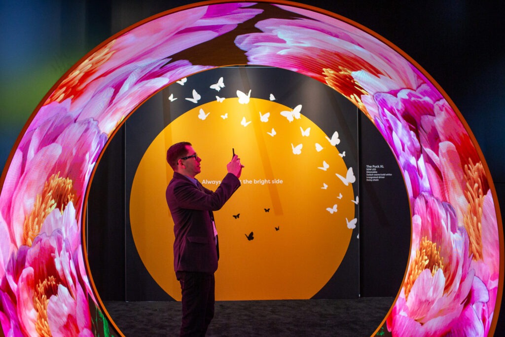 Gebogene LED-Installation als leuchtender Ring mit Blumenmotiven; Besucher fotografiert mit Smartphone, dahinter runde orange Displayfläche mit weißen Schmetterlingen