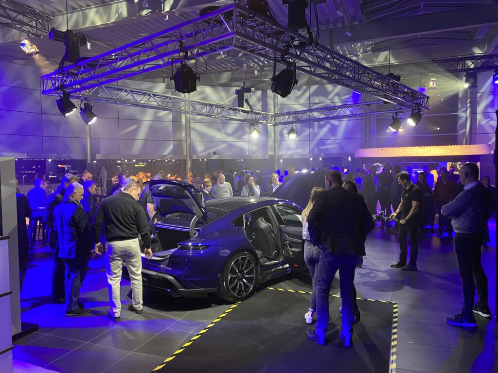 Autohaus-Event: Sportwagen-Präsentation mit geöffneten Türen, zahlreiche Gäste, Traversengrid mit bewegtem Blau-/Violettlicht, Kamerateam und Bar im Hintergrund