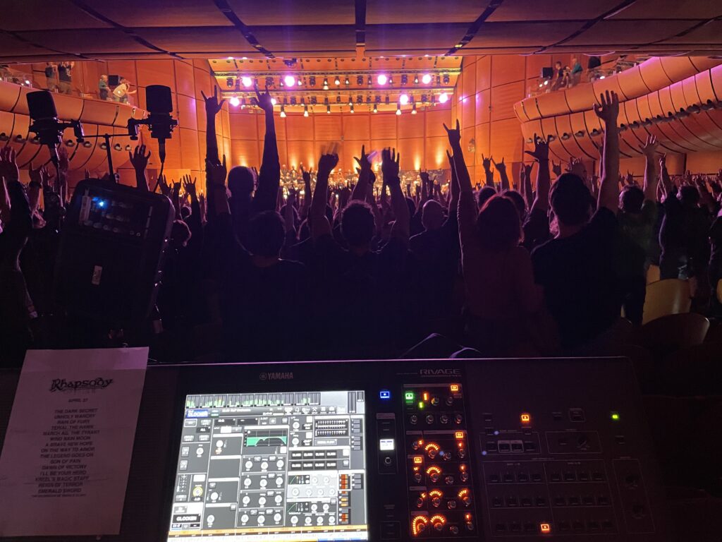 Blick vom Technikpult aus in einen Konzertsaal voller jubelnder Zuschauer mit erhobenen Händen, während auf der hell erleuchteten Bühne eine Live-Performance stattfindet. Im Vordergrund ist ein Yamaha-Rivage-Mischpult zu sehen, daneben eine Setlist mit dem Logo der Band "Rhapsody of Fire".