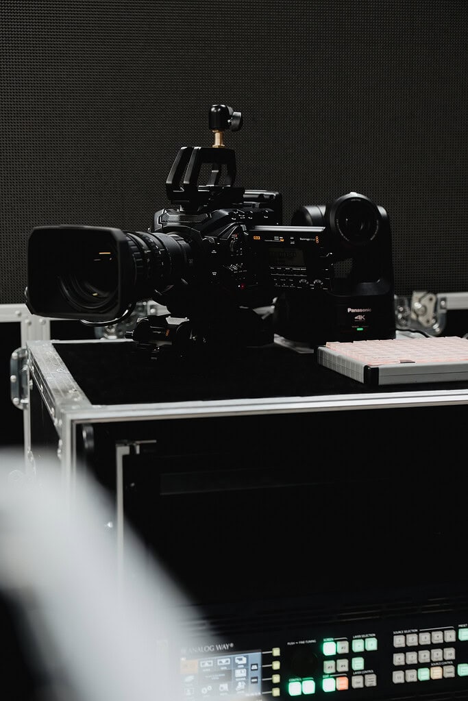 Professionelle Videokamera auf Technik-Case; daneben PTZ-Kamera und Videomischer im Bühnen-Backstage.